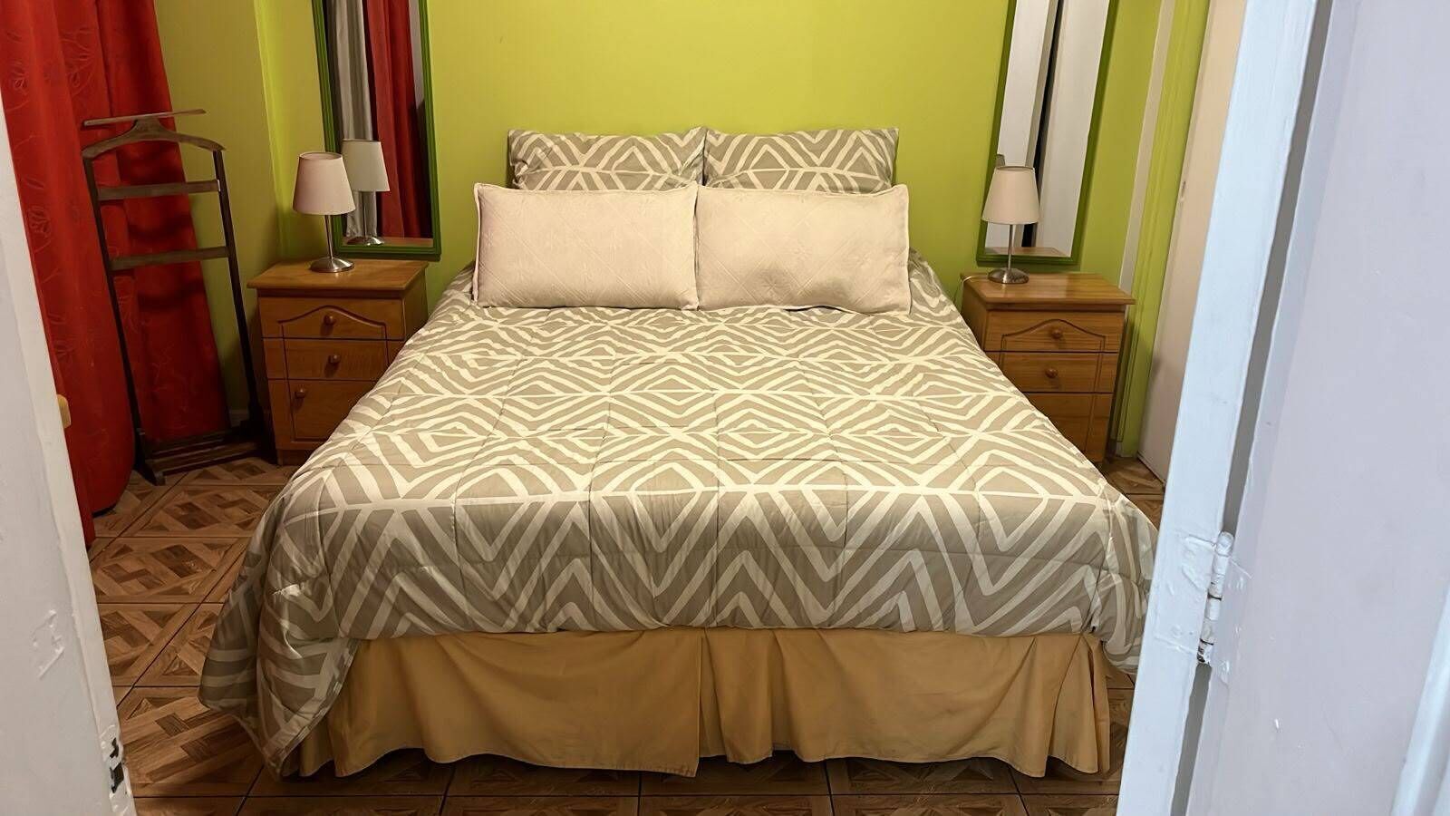 Una cama con colcha estampada, dos mesitas de noche con lámparas, una silla de madera y una cortina roja.