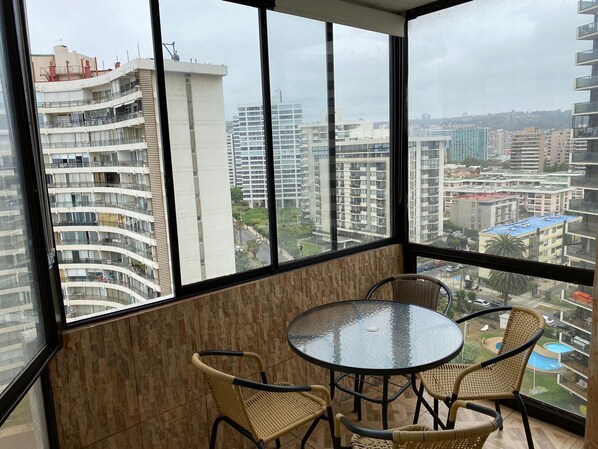 A balcony with a round glass table and wicker chairs, overlooking a cityscape with high-rise buildings.