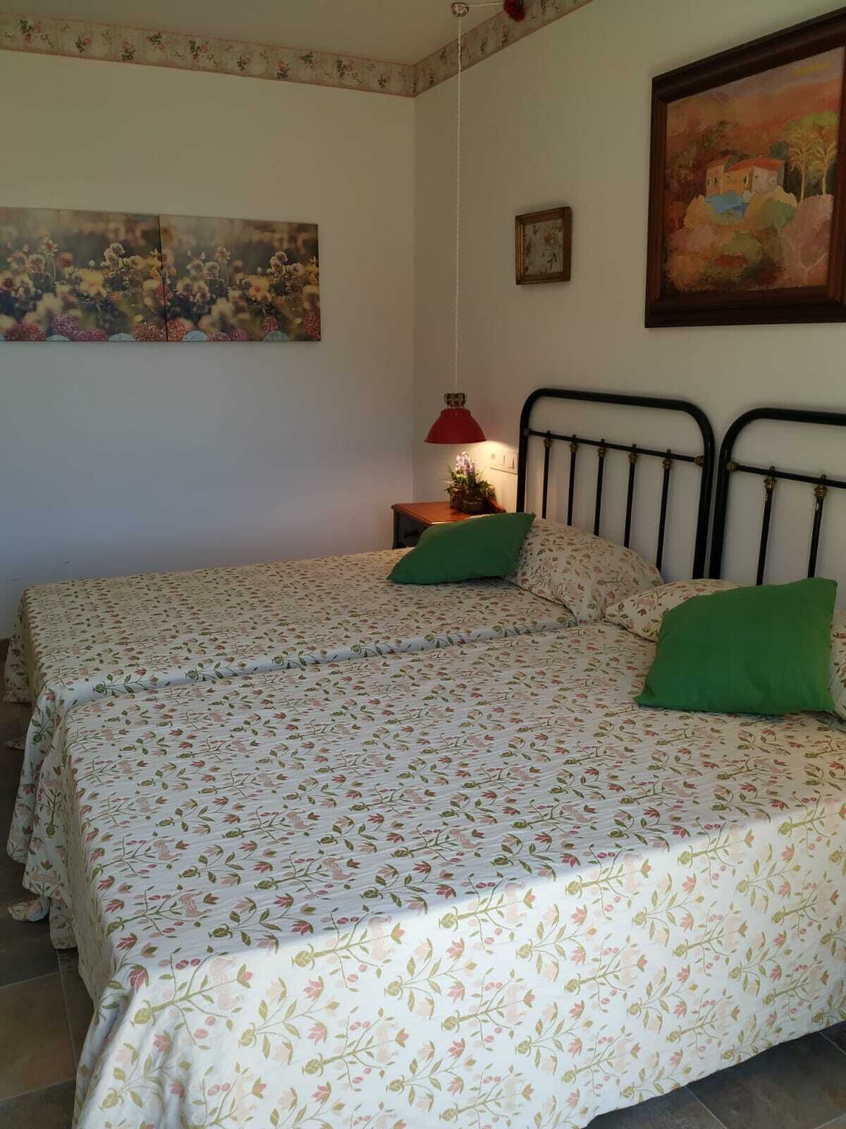 A room with two beds, each with floral-patterned bedding, a red lamp on a bedside table, and a painting on the wall.