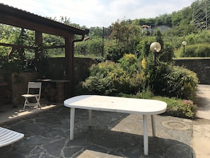 A patio with a white table and chairs, surrounded by stone flooring and lush greenery. - Villa surrounded by greenery overlooking Lake Maggiore (Pisano)