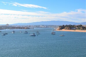 A coastal view with multiple sailboats on calm waters, a sandy beach, and distant mountains.