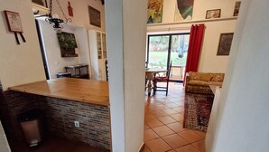 Een keuken met een houten aanrecht en een betegelde achterwand, een eethoek met een tafel en stoelen, en een woonkamer met een bank en een vloerkleed met een patroon.