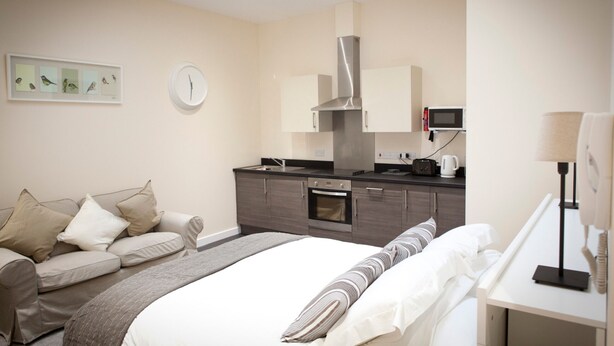 A modern hotel room with a bed, a sofa, a kitchenette, and a clock on the wall. - Toothbrush Apartments - Central Ipswich East - Rope Walk (Ipswich)