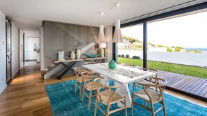 Un comedor moderno con una mesa blanca y sillas de madera, una alfombra azul y vistas a una masa de agua a través de amplios ventanales.
