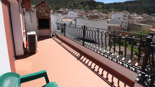 A balcony with a metal railing, a small building with a chimney, and a view of a town with buildings and hills in the background.