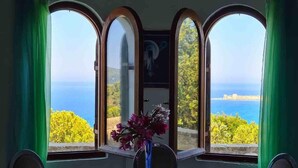 A dining area with a table and chairs, a vase with flowers, and a view of the sea through arched windows.