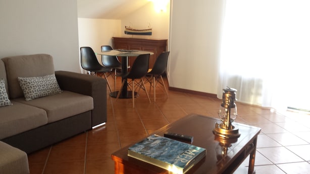 A living room with a sofa, a coffee table with a book and a decorative item, a dining area with chairs and a table, and a kitchen in the background.