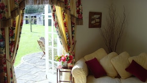 A living room with a beige sofa, a small table with a vase of flowers, and a view of a garden through a glass door.