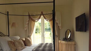 A four-poster bed with a white bedspread and multiple pillows, a bedside table with a mirror, and a view of a garden through a large window.