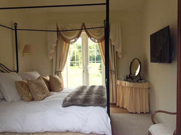 A four-poster bed with a white bedspread and multiple pillows, a bedside table with a mirror, and a view of a garden through a large window.