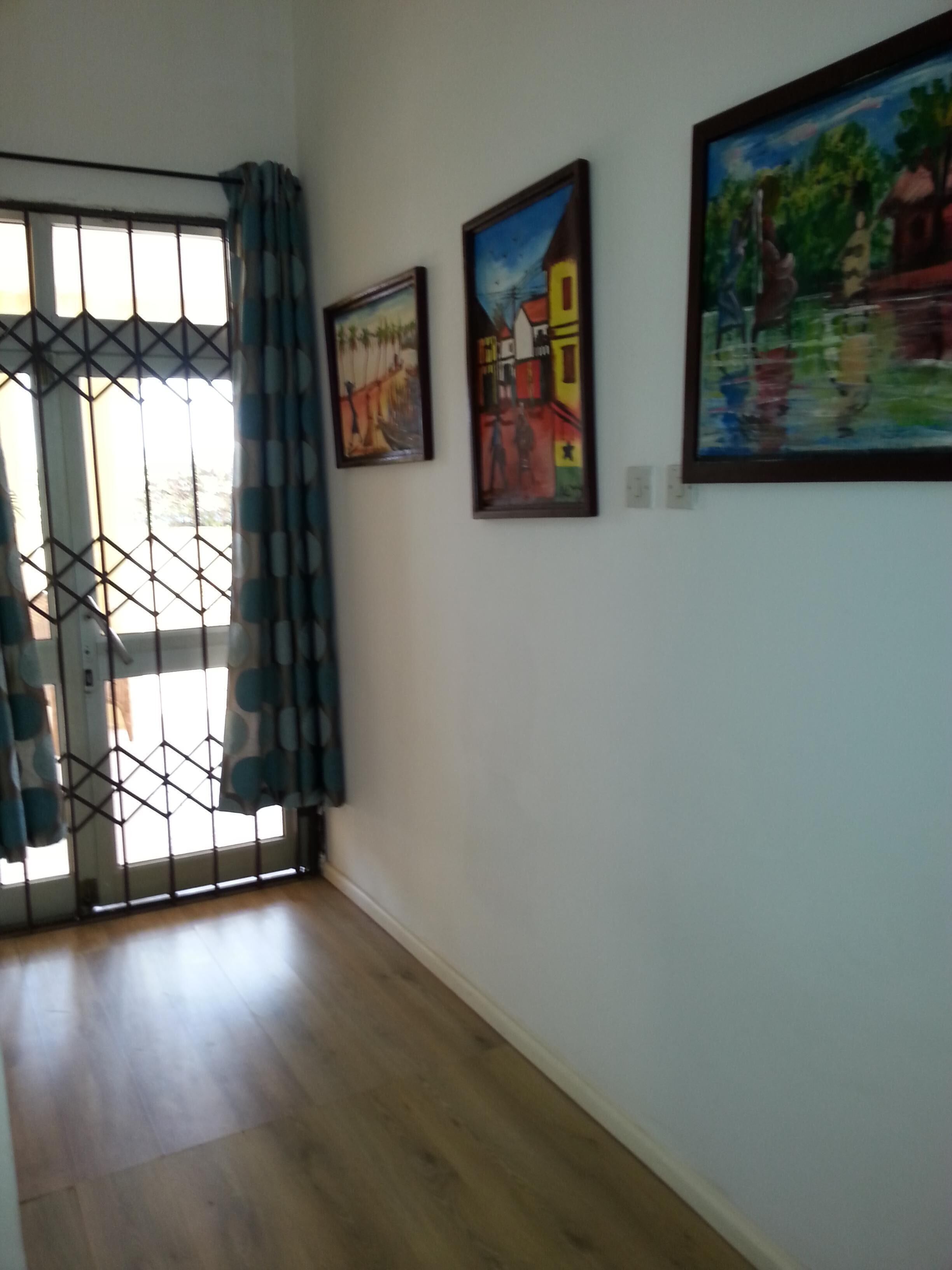 A room with wooden flooring, a window with metal bars, and three framed paintings on the wall.