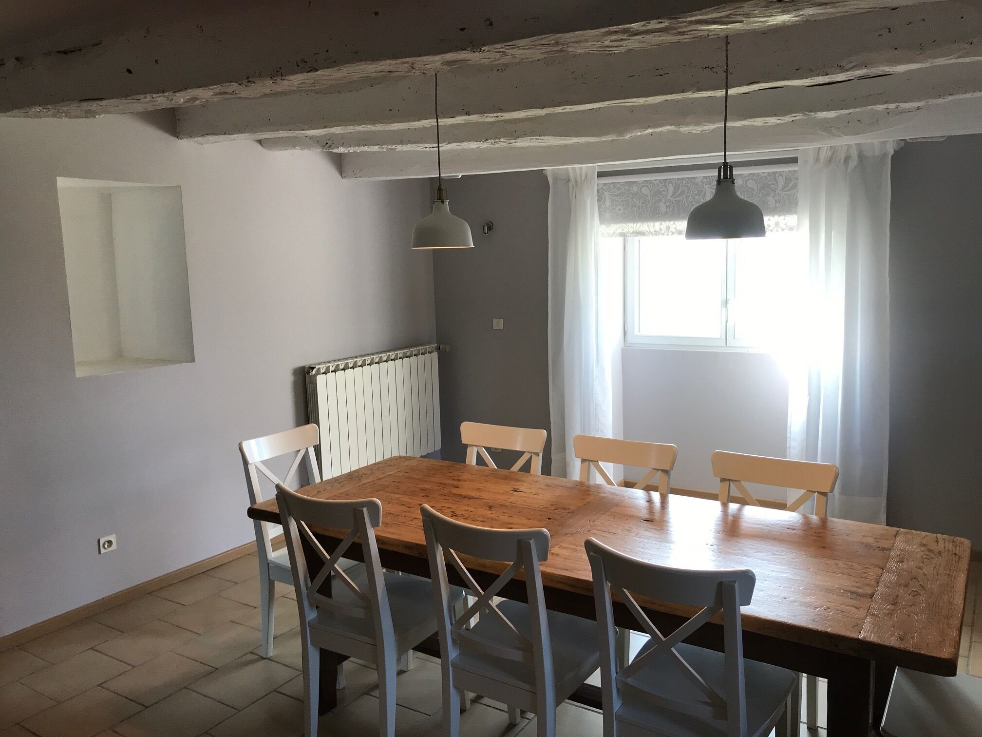 Une salle à manger avec une table en bois et des chaises blanches, un radiateur, un luminaire suspendu et une fenêtre avec des rideaux.