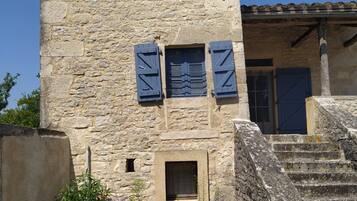 Un edificio de piedra con una torre, un ventanal con contraventanas y una escalera que conduce a una puerta.