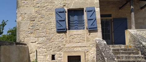 A stone building with a tower, a small window with shutters, and a staircase leading to a door.