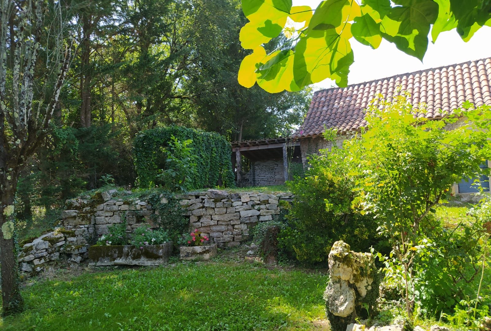 Un muro de piedra, un edificio rústico con techo de tejas y abundante vegetación.