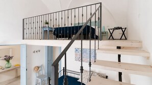 Eine Treppe mit Metallgeländer führt zu einem Schlafzimmer auf einem Zwischenstock, das ein Bett und einen Schreibtisch beinhaltet.