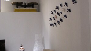 Una sala de estar con un sofá color beige, un adorno de pared con pájaros, una lámpara blanca y una mesita con un jarrón.