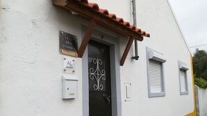 Un edificio con fachada blanca y amarilla, una puerta negra con detalles de metal decorativo y una ventana pequeña con contraventanas.
