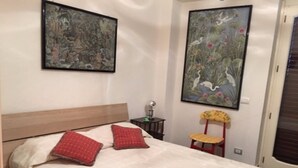A bedroom with a wooden headboard, two red pillows, a bed with a white quilt, a small table with a lamp, and two framed artworks on the wall. - Apartment in villa with air-conditioned pool (Castelbuono (Pa))