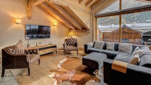 Een gezellige woonkamer met een houten plafond, een groot raam, een leren bank, een fauteuil bekleed met leer en een koperen vloerkleed.