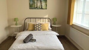 A neatly made bed with white bedding and a rolled towel, two green table lamps on side tables, a painting of a forest on the wall, and a window with curtains. - Cosy Corner House. (Kettering)
