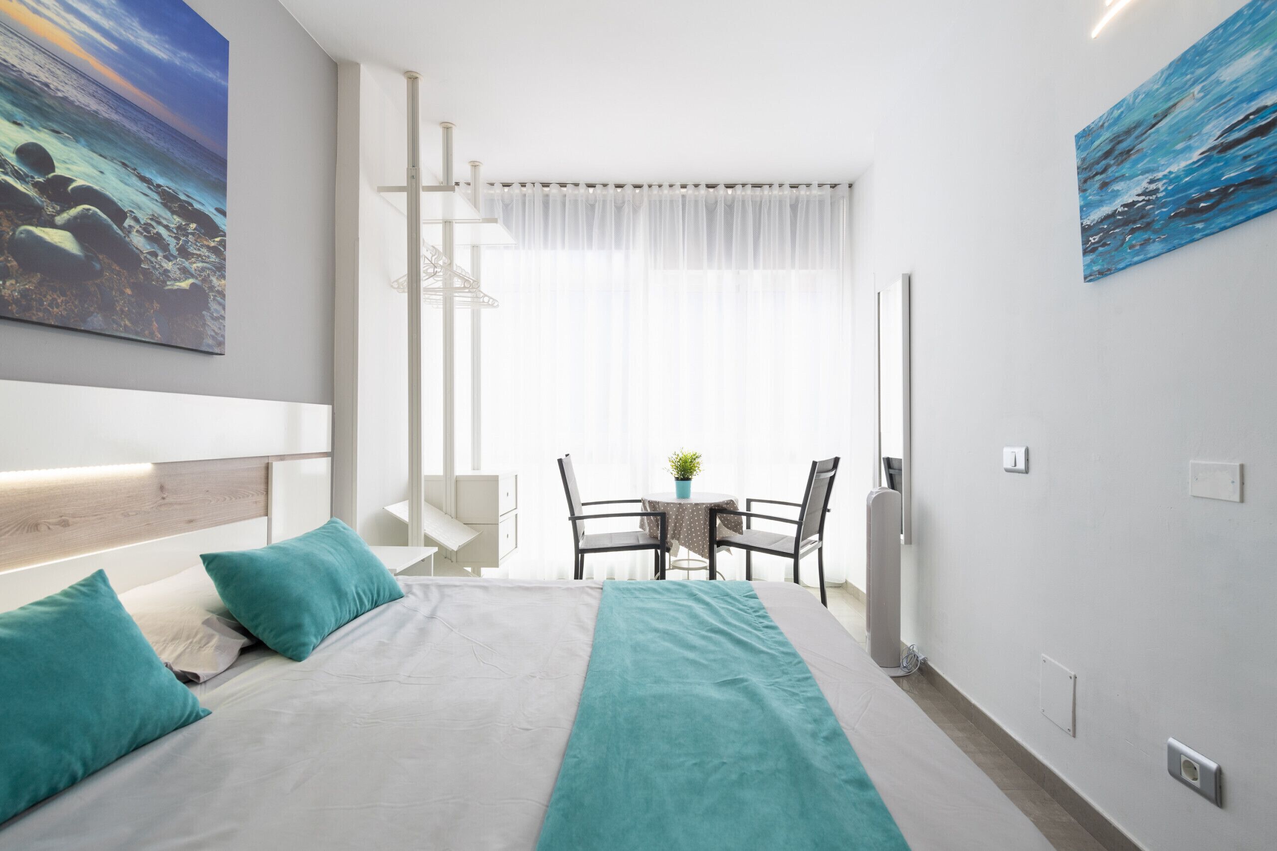 A modern hotel room with a bed, two chairs, a small table, and a view of the sea through sheer curtains.