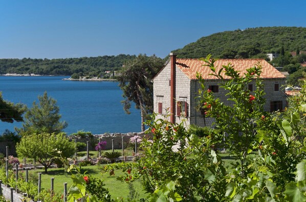A stone building with a red-tiled roof near a body of water, surrounded by greenery and a well-maintained garden.