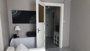 Een moderne hotelkamer met een witte bank, een abstract schilderij in het zwart, een tafeltje met een lamp, en een deur die uitkomt op een gang.