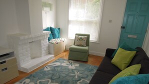A living room with a fireplace, a green armchair, a black sofa with colorful cushions, and a patterned rug. - Pretty Victorian cottage (1860) in a charming setting, 15 min walk from seafront (Portsmouth)