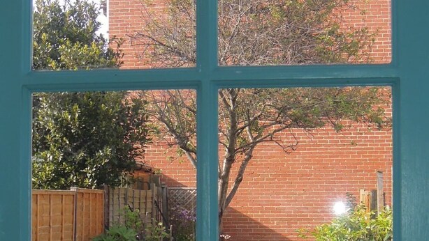 A view through a window with a brick building, a wooden fence, and a garden with flowers and shrubs. - Pretty Victorian cottage (1860) in a charming setting, 15 min walk from seafront (Portsmouth)