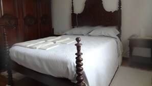 Una cama con dosel de madera, con cubrecama y almohadas blancas, una mesita de noche de madera con lámpara, y un armario con espejo.
