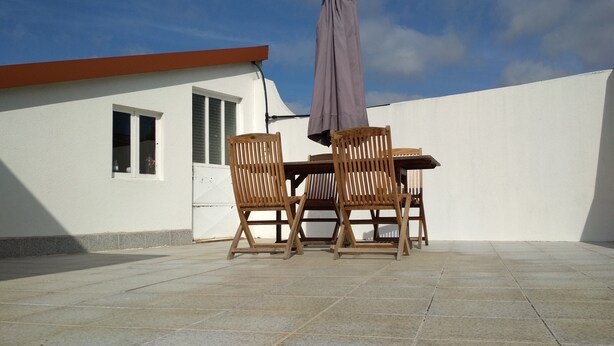 Un patio al aire libre con una mesa, dos sillas y una sombrilla, con un edificio blanco de techo rojo al fondo.