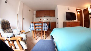 Habitación pequeña con cama, una silla, mesa de comedor con sillas, televisor y zona de cocina.