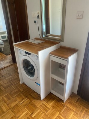 A compact, white washing machine with a wooden top and a small shelf, placed in a corner of a room with wooden flooring and a mirror above it. - Peaceful holidays for an active family (Stari Grad)