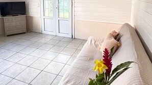 A spacious room with a high ceiling, a sofa with a cushion, a small table with a vase of flowers, and a television on a cabinet. - 🌴 Creole house in the heart of the Martinique countryside (Gros-Morne)