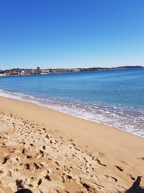 Ein sandiger Strand mit Fußspuren, die zum Wasser führen, ein klarer, blauer Himmel und eine ferne Stadtlandschaft.