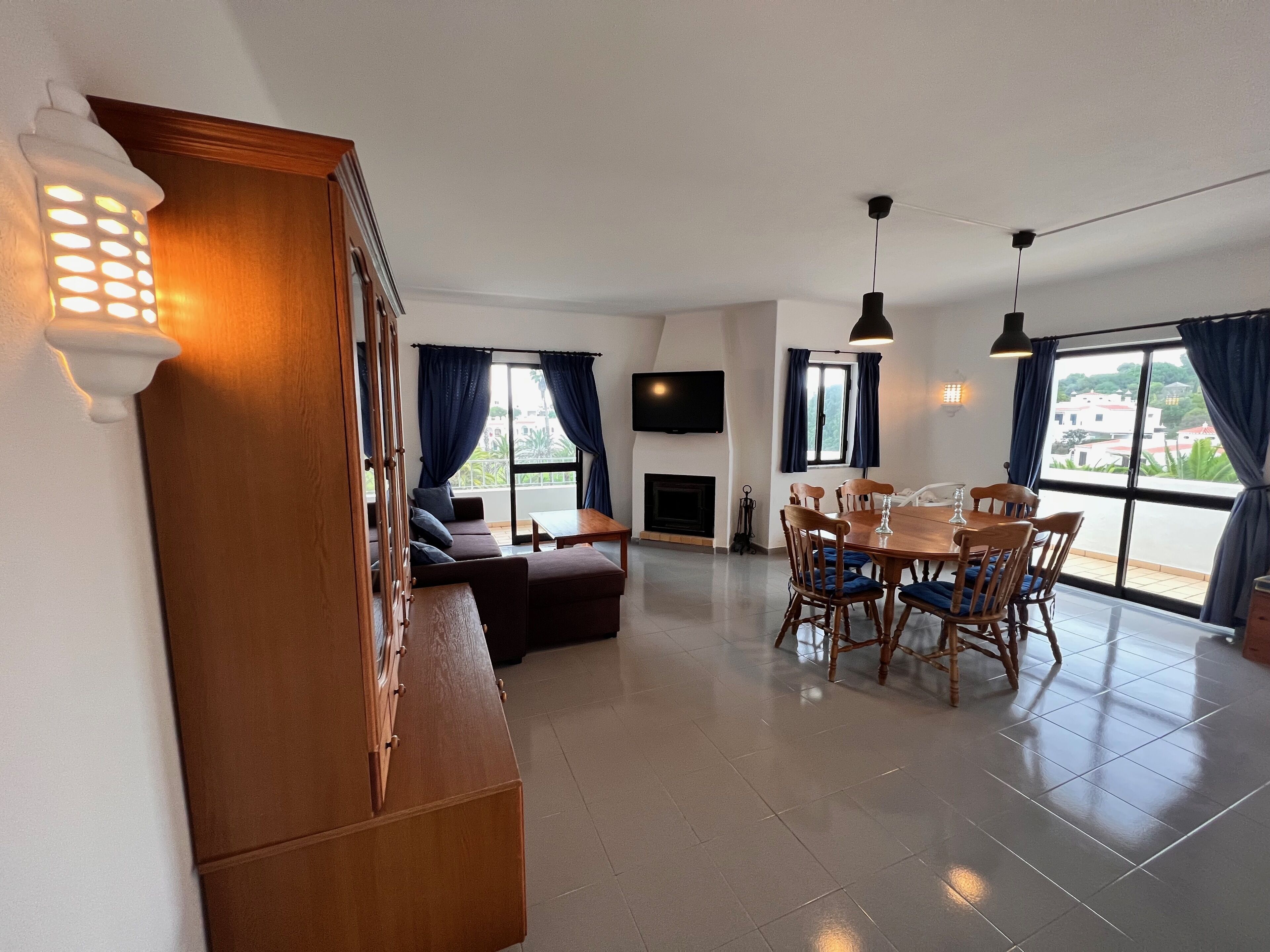 A modern living room with a sofa, dining table, and chairs, a fireplace, and large windows with curtains.