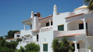 Un bâtiment blanc doté de nombreux balcons et de volets verts, entouré de verdure.