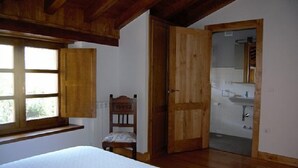 Quarto com teto de madeira, janela, cadeira e porta que dá para um banheiro.