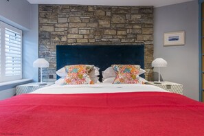 Un dormitorio con pared de piedra, cabecera oscura, colcha roja y dos lámparas de noche.