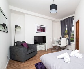 Una habitación de hotel moderna con sofá gris, televisor de pantalla plana, zona de comedor con mesa y sillas, y una cama grande con toallas.