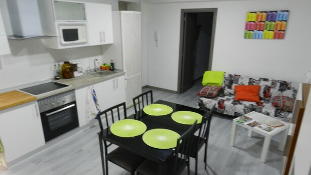 Una cocina funcional con armarios blancos, microondas y estufa. Hay una mesa de comedor con cuatro sillas y una mesita auxiliar con revistas.