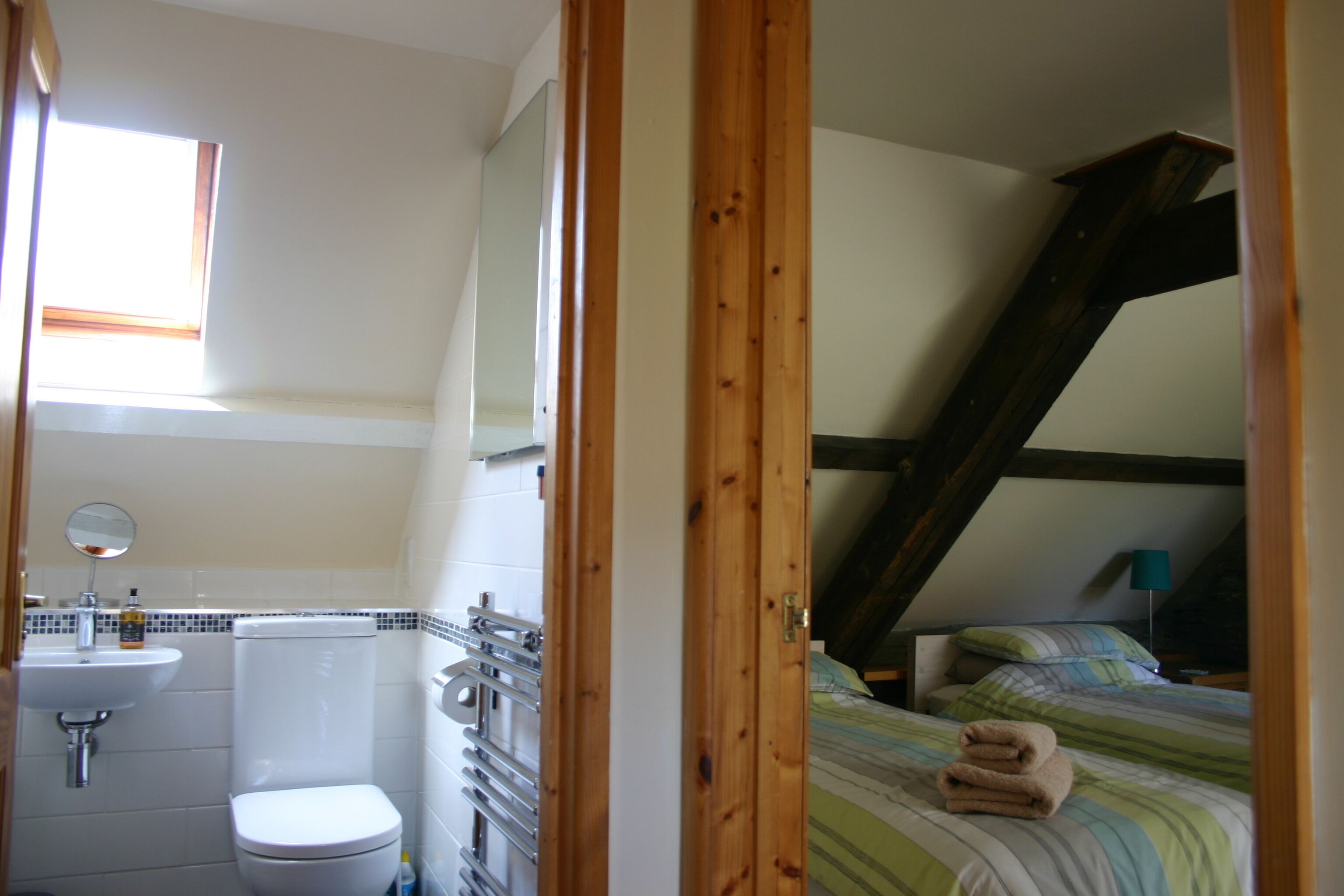 Een badkamer met toilet, wastafel en spiegel. Er is een raam boven de wastafel en een bed met een gestreepte deken in de aangrenzende kamer.