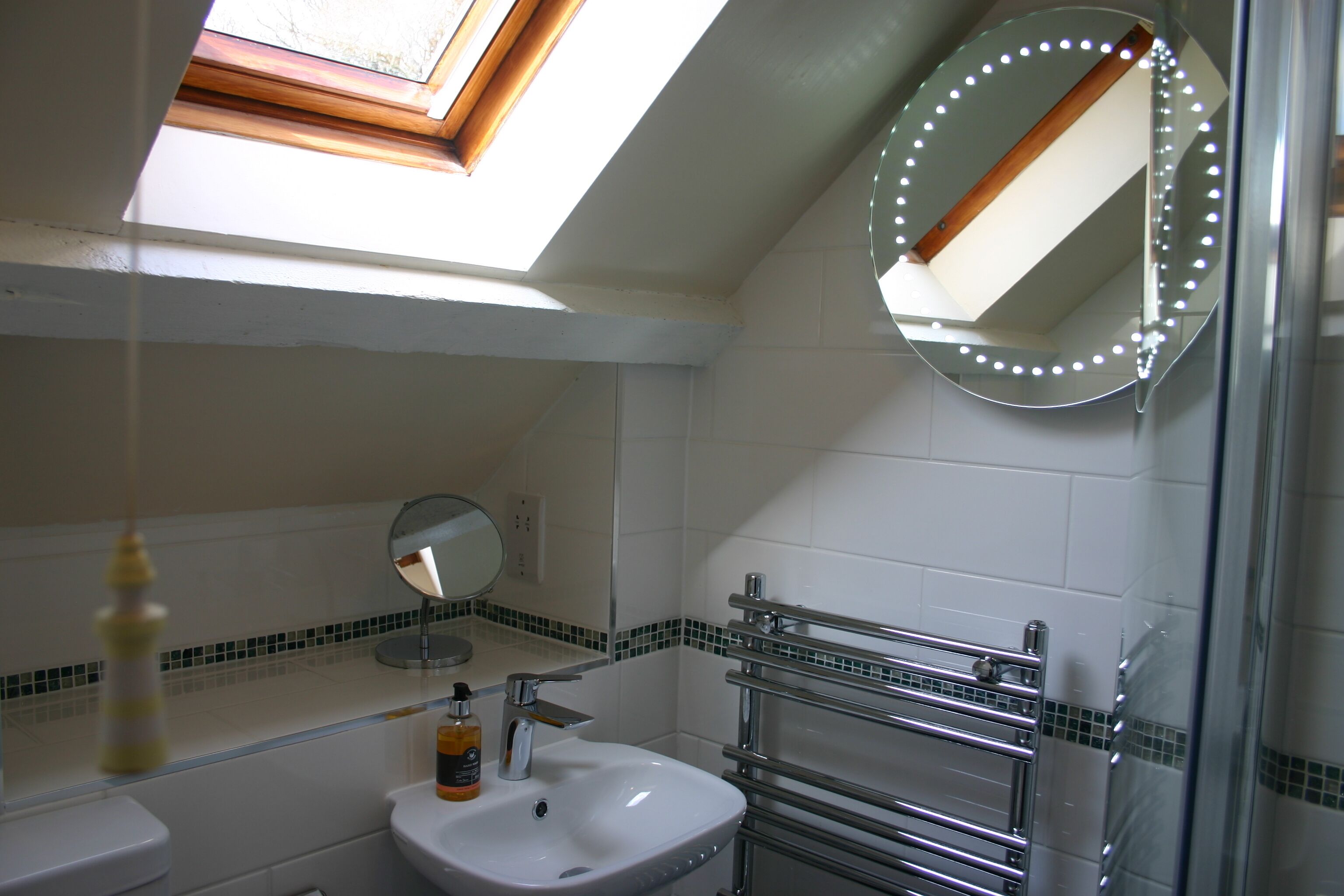 A bathroom with a skylight, a round mirror with lights, a sink, and a towel rack.