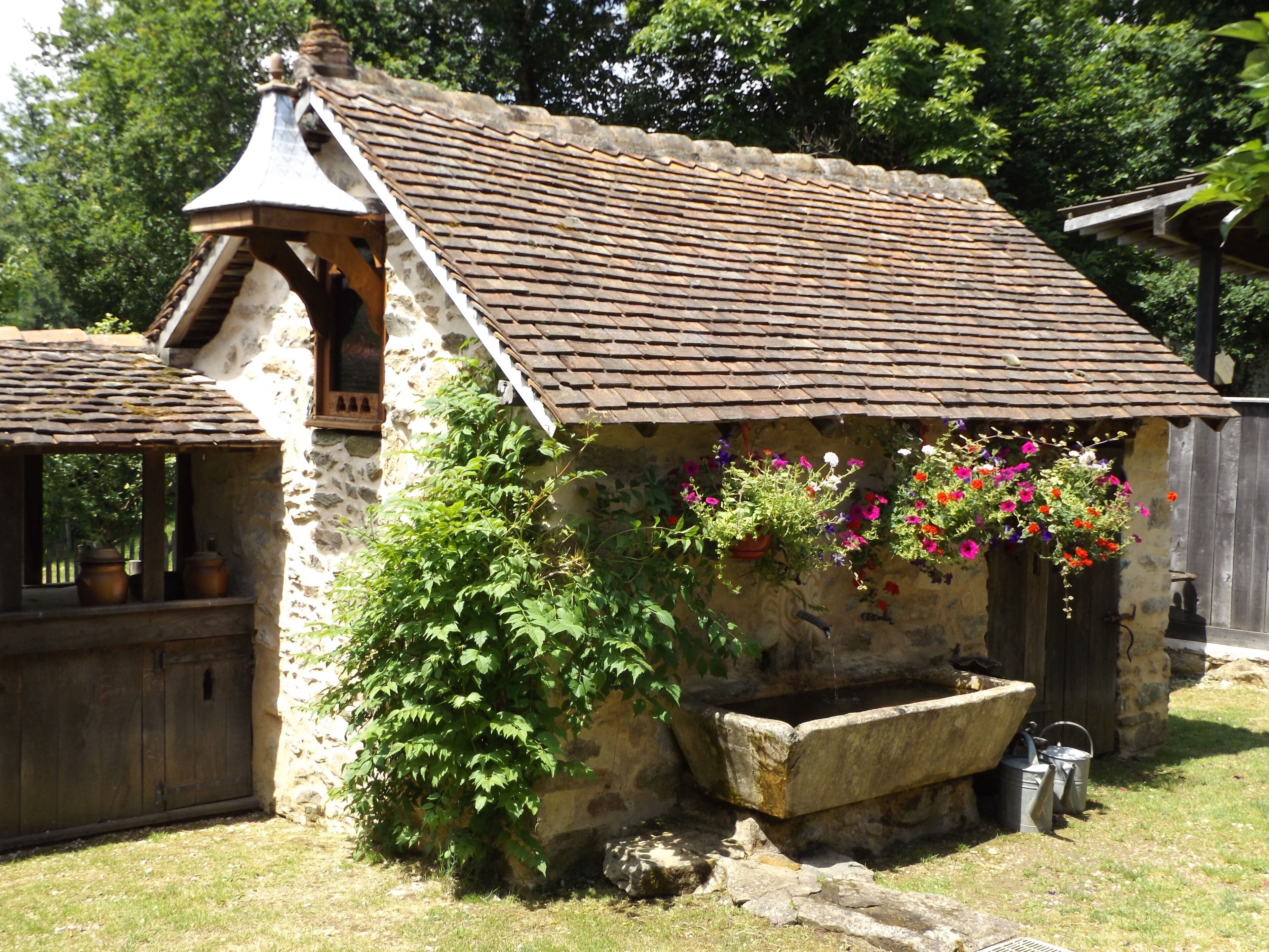 Un edificio rústico de piedra con techo inclinado, puerta de madera y una ventana. Hay plantas en macetas y cestas de flores colgantes en el exterior.