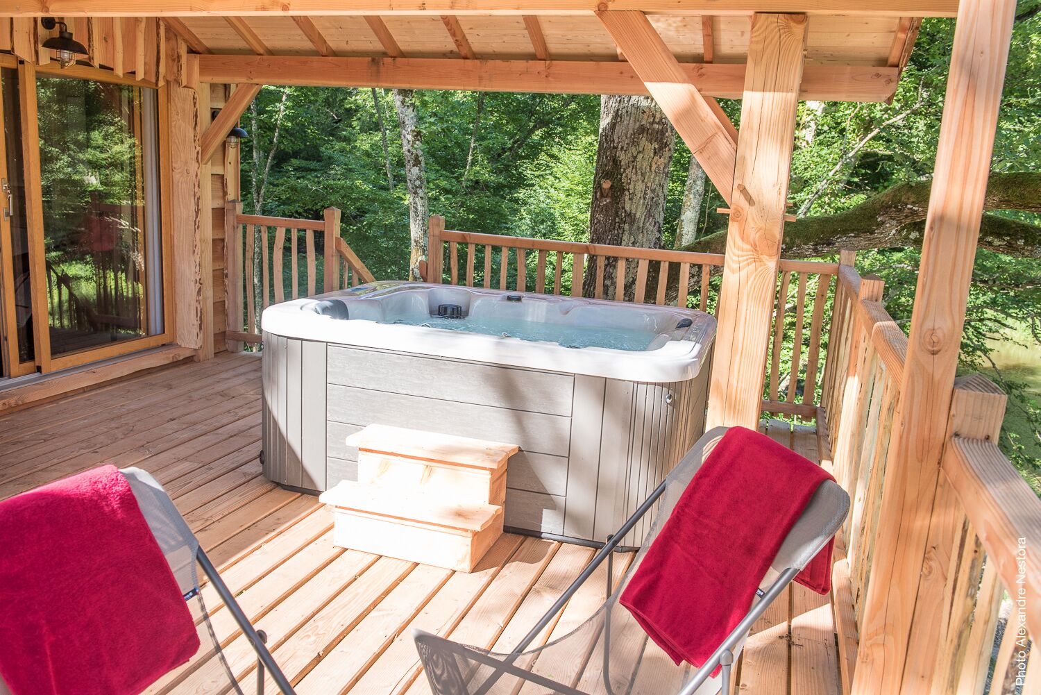 Une terrasse en bois avec un jacuzzi, deux chaises accompagnées de serviettes rouges et une porte vitrée donnant sur une zone boisée.