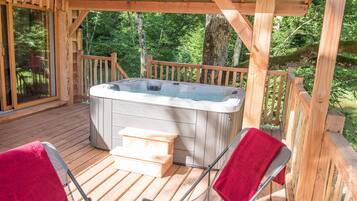 Une terrasse en bois avec un jacuzzi, deux chaises accompagnées de serviettes rouges et une porte vitrée donnant sur une zone boisée.