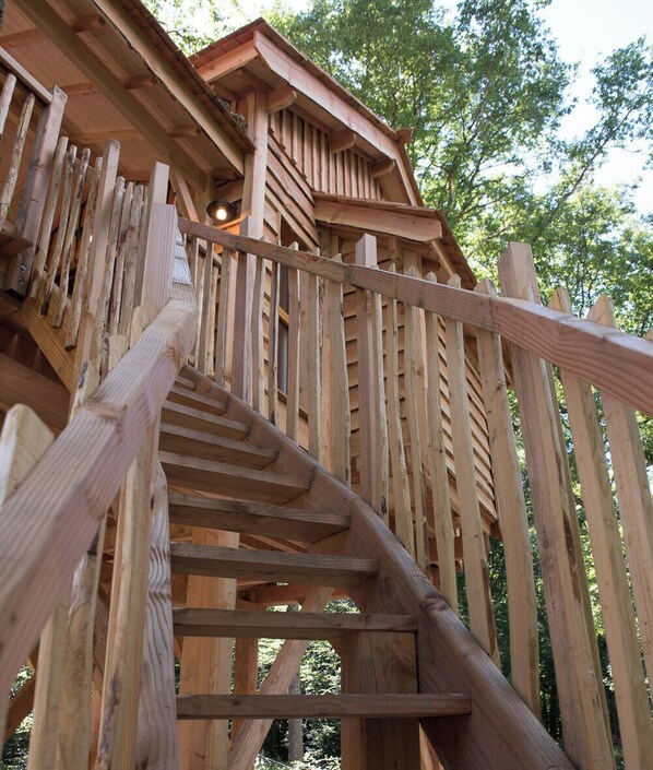 A wooden spiral staircase leading to a wooden structure with a sloped roof, surrounded by trees.