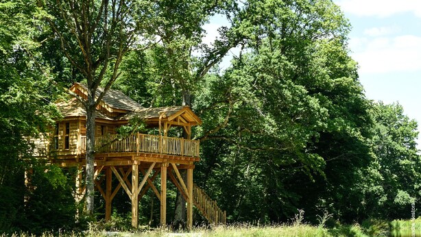 A wooden treehouse with a balcony, situated by a river surrounded by dense greenery. - LUXURY CABIN WITH SPA IN TREES (La Bussiere)
