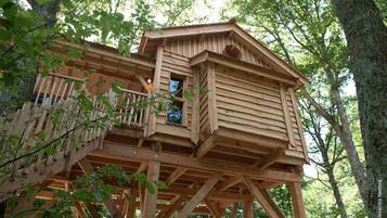 Une cabane en bois perchée dans un arbre, dotée d’un balcon et construite sur pilotis, entourée d’arbres.